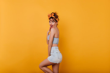 Surprised shapely girl covering mouth with hand and laughing. Lovable european woman in light-blue shorts posing emotionally on yellow background.