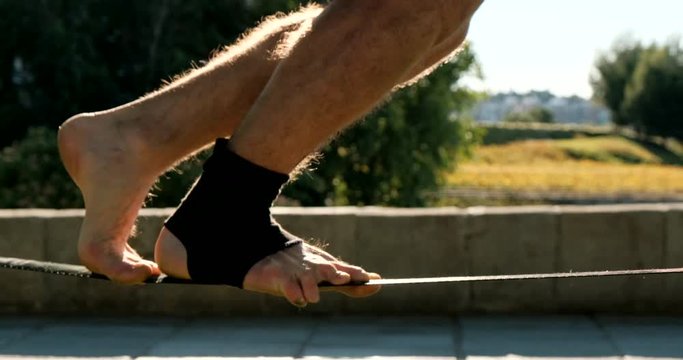 Close-up of male feet walking on rope. Young man performing as acrobat and training with slackline outdoors. Athlete doing sport activity and exercising with trickline in city park