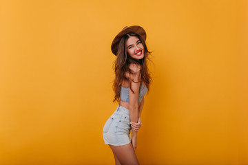 Fascinating girl with shy smile expressing positive emotions. Indoor portrait of good-looking young woman with dark hair smiling on yellow background.