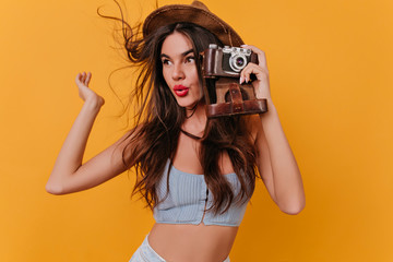 Glad girl with long hairstyle holding camera and waving hand. Studio shot of female photographer in...