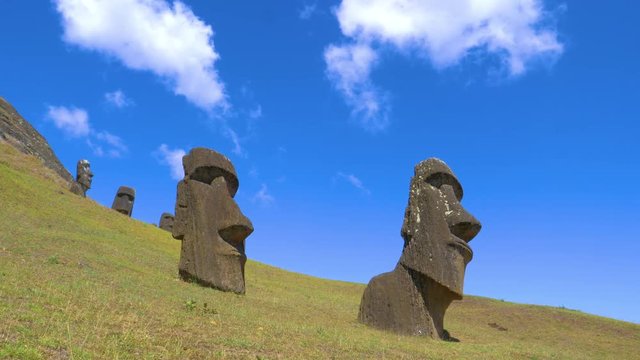 LOW ANGLE: Beautiful blue summer sky with puffy white clouds spans above a group of ancient moai sculptures. Cool shot of scenic Easter Island landscape filled with statues made of black volcanic rock