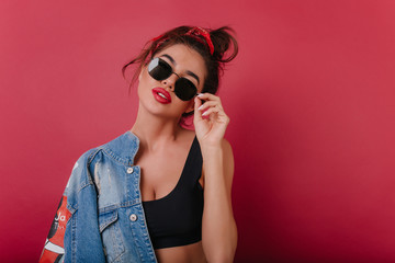 Slim girl with red lips standing on dark background and touching black sunglasses. Studio photo of elegant white female model with short hairstyle holding jeans jacket.
