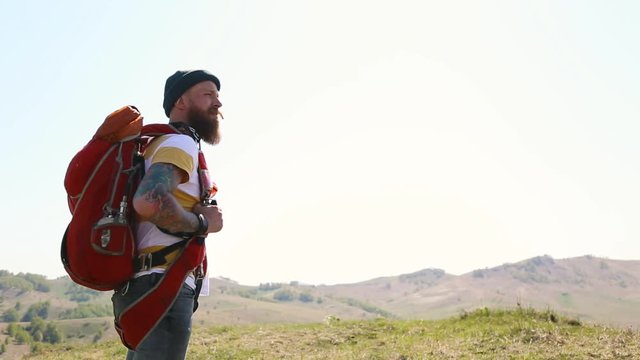 man with beard mustache with red backpack parachute green hat striped white yellow T-shirt jeans, walks through mountains summer and looks around. tattoo on hand. caucasian