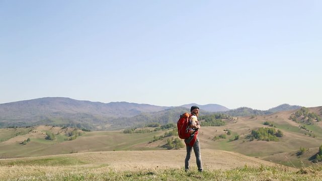 man with beard mustache with red backpack parachute green hat striped white yellow T-shirt jeans, walks through mountains summer and looks around. tattoo on hand. caucasian