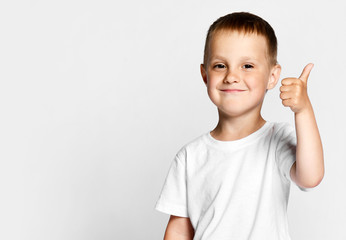 Little cute boy preschooler 4 years old in a white T-shirt and gray pants on a light background, happily looking at the camera, showing a thumb sign cool