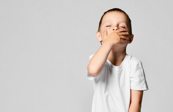 Child Covers His Mouth With His Hand And Yawns His Eyes Closed. Portrait Of A Young Boy Isolated On White Background