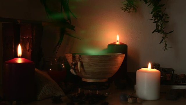 Detail of a potion making witch room, with green steam coming out from a wooden bowl, animal teeth hanging from it, candles with flickering flames, old books, glasses and herbs in the background