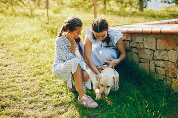 two beautiful and cute girls in blue dresses with beautiful hairstyles and make-up sitting in a sunny green garden and playing with a dog
