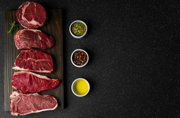 Selection of raw beef meat food steaks against black stone background. New york striploin steak, top blade, rib eye, and other cuts of meat.