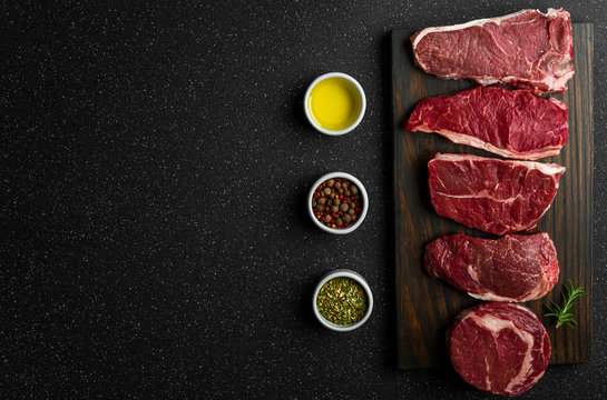 Selection of raw beef meat food steaks against black stone background. New york striploin steak, top blade, rib eye, and other cuts of meat.