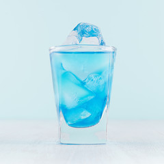 Tropical fresh alcohol cocktail with blue curacao liqueur, ice cube in frozen shot glass on soft light white, pastel green background.