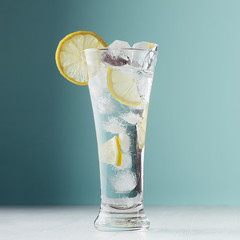 Fresh summer citrus cold drink with yellow lemon pieces, ice cubes, tonic in light soft pastel ...