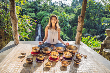 Woman playing on Tibetan singing bowl while sitting on yoga mat against a waterfall. Vintage tonned. Beautiful girl with mala beads meditating