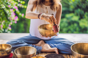 Woman playing on Tibetan singing bowl while sitting on yoga mat against a waterfall. Vintage tonned. Beautiful girl with mala beads meditating