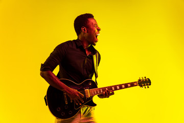 Young african-american musician playing the guitar like a rockstar on yellow background in neon light. Concept of music, hobby, festival, open-air. Joyful attractive guy improvising, singing song.