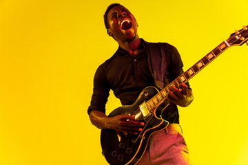 Young african-american musician playing the guitar like a rockstar on yellow background in neon light. Concept of music, hobby, festival, open-air. Joyful attractive guy improvising, singing song.