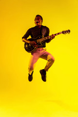 Young african-american musician playing the guitar like a rockstar on yellow background in neon...