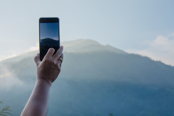 Womans hand taking photo of mouintain in late afternoon with phone.