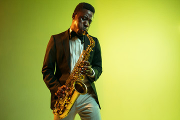 Young african-american jazz musician playing the saxophone on gradient yellow-green studio...