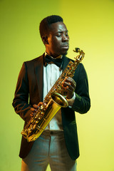 Young african-american jazz musician playing the saxophone on gradient yellow-green studio...
