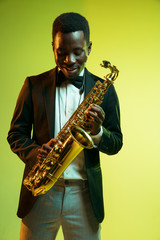 Fototapeta na wymiar Young african-american jazz musician playing the saxophone on gradient yellow-green studio background. Concept of music, hobby, festival. Joyful attractive guy improvising. Colorful portrait of artist