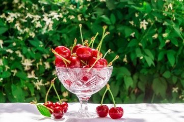 Fresh cherries in crystal bowl on a table with a white cloth on a green natural background of blurred flowers. Concept of healthy food, vitamin C. Copy space for your text