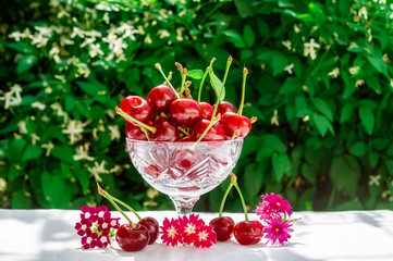 Fresh cherries in crystal bowl on a table with a white cloth and decor of flowers on a green natural background of blurred flowers. Concept of healthy food, vitamin C. Copy space for your text