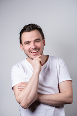 Handsome brunette man in a white T-shirt with arms crossed is smiling on a gray background