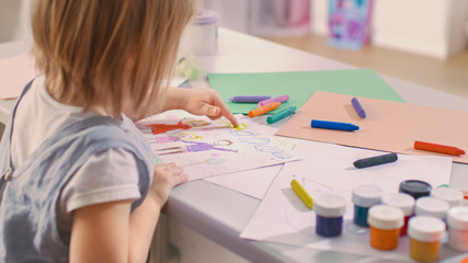 Back Shot of a Cute Little Girl Sits at Her Table and Draws. She Paints Sun with Her Yellow Finger. Her Room Is Pink and Cosy.