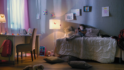 Cute Little Girl Lies on Her Bed and Watches Cartoons on a Smartphone. Her Kitten is Beside Her Playing. Her Floor Lamp is On.