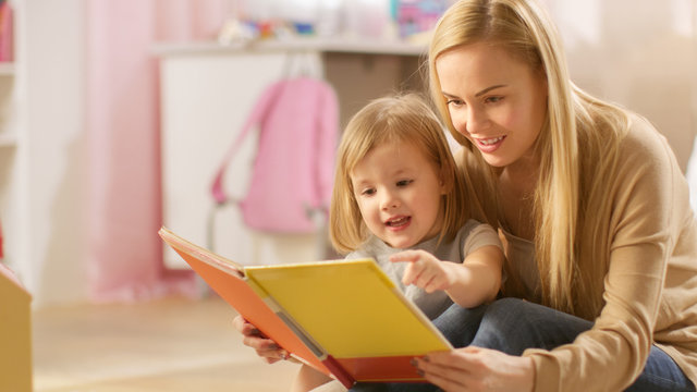 Beautiful Young Mother and Her Cute Little Daughter Read Children's Book Together. Children's Room is Pink and Full of Toys.