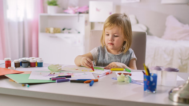 Cute Little Girl Sits at Her Table and Draws with Crayons. Her Room Is Pink, Pretty Drawings Hanging on the Walls, Many Toys Lying Around.