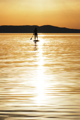 Centered sun reflection in water and silhouette of young woman paddling at sunset on a stand up paddleboard (SUP) in Croatia, Adriatic Sea, near Sibenik. Vertical photo.