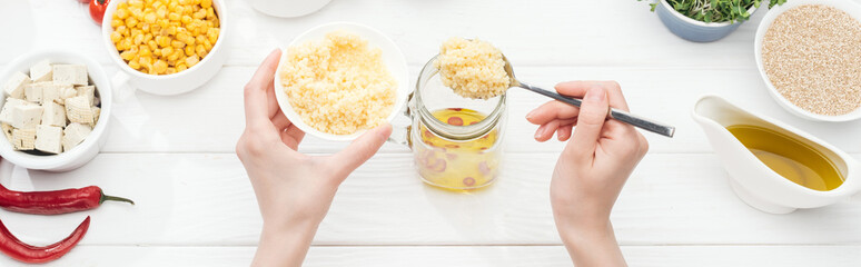 partial view of woman adding couscous to glass jar on wooden white table, panoramic shot