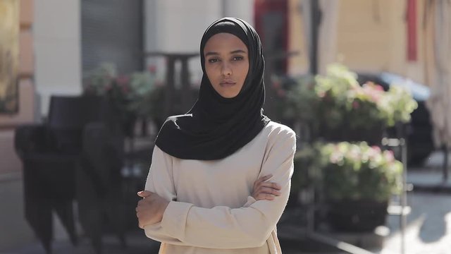 Portrait of beautiful young Muslim woman wearing hijab headscarf looking into the camera standing with crossed hands on the old city background.