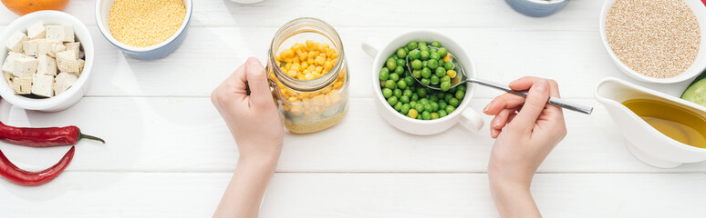 cropped view of woman adding green peas in jar with salad on wooden white table, panoramic shot