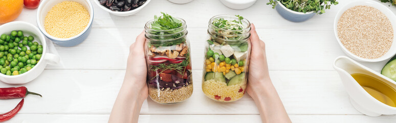 cropped view of woman holding jars with tasty salad on wooden white table , panoramic shot