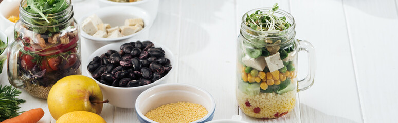 ingredients in bowl near glass jars with salad on wooden white table, panoramic shot