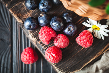 Raspberries and blueberries in a basket with chamomile and leaves on a dark background. Summer and healthy food concept. Selective focus. Flat lay.