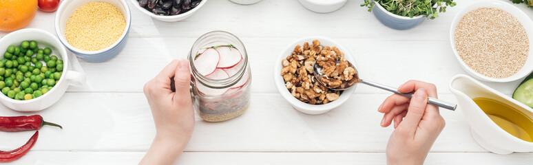 cropped view of woman adding nuts in jar with salad on wooden white table, panoramic shot