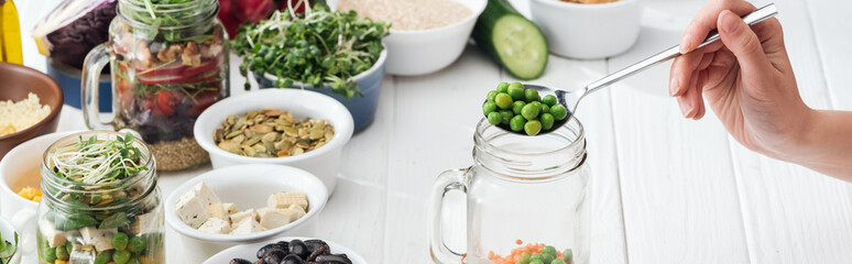 cropped view of woman adding green peas in glass jar on wooden white table, panoramic shot