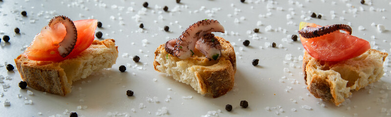 Top view of tasty Italian appetizers -  bruschetta with raw tomatoes and octopus tentacles, on slices of toasted baguette. White background with black pepper and sea salt.