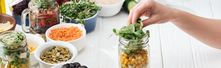 cropped view of woman adding greens in glass jar on wooden white table, panoramic shot