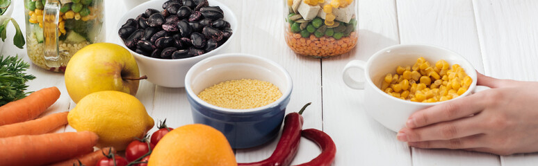 cropped view of woman holding corn in bowl on wooden white table, panoramic shot