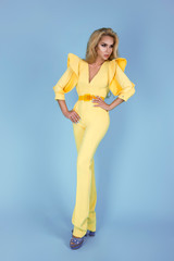 Elegant blonde woman in stylish yellow jumpsuit and fashionable accessories on color background. Beautiful fashion model on blue background.