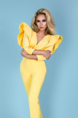 Elegant blonde woman in stylish yellow jumpsuit and fashionable accessories on color background. Beautiful fashion model on blue background.