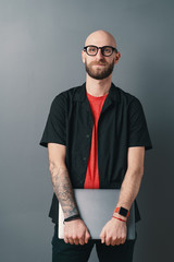 Responsible bearded man with glasses holding laptop under arm in the studio on gray background