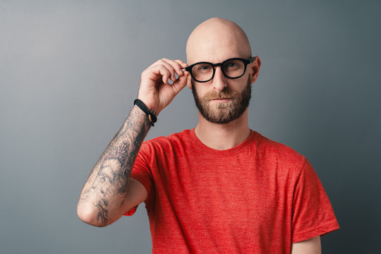 Confident young bearded man in red t-shirt fixing his black framed glasses on gray background
