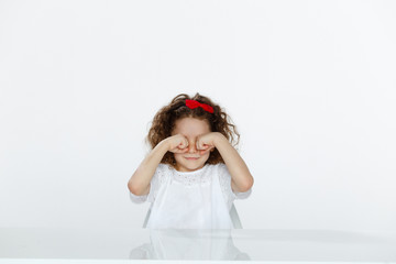 Adorable curly little girl in studio, seated at a table, she's rubbing his eyes with hands, isolated on a white backgrounds.