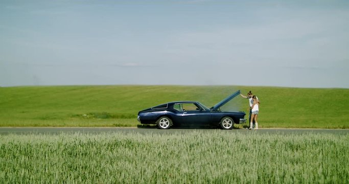 Couple standing near overheated car in the field, bright sunlight, smoke from under the hood. 4K UHD RAW graded footage
